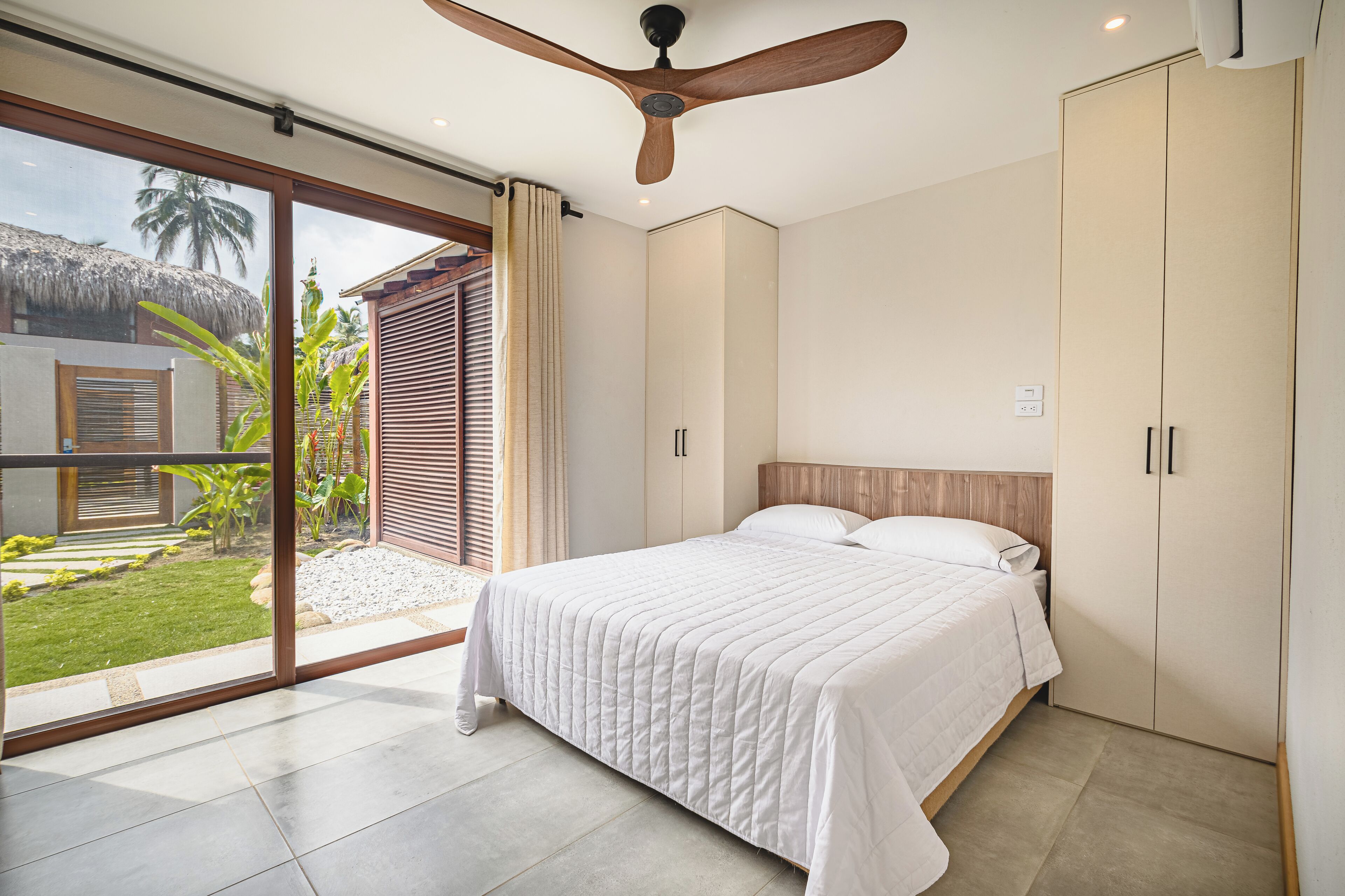 A bedroom with a large bed, sliding glass doors to a garden, a ceiling fan, and a wardrobe.