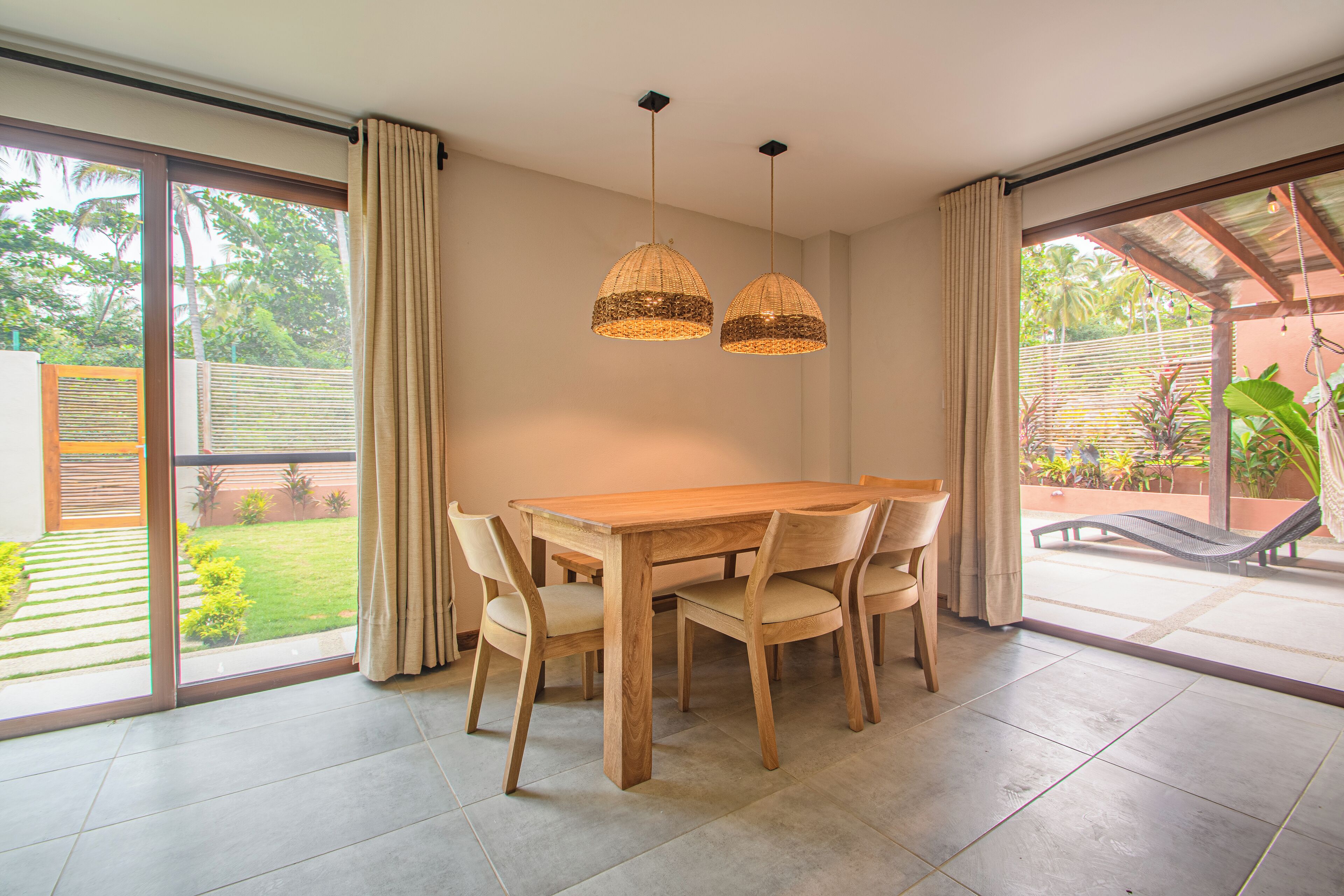 Un comedor con mesa y sillas de madera, dos lámparas colgantes y vistas a un jardín a través de puertas correderas amplias.