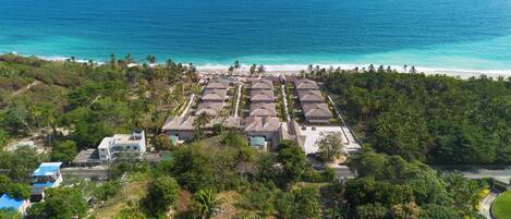 Un resort con edificios de techo de paja, una playa y abundante vegetación.