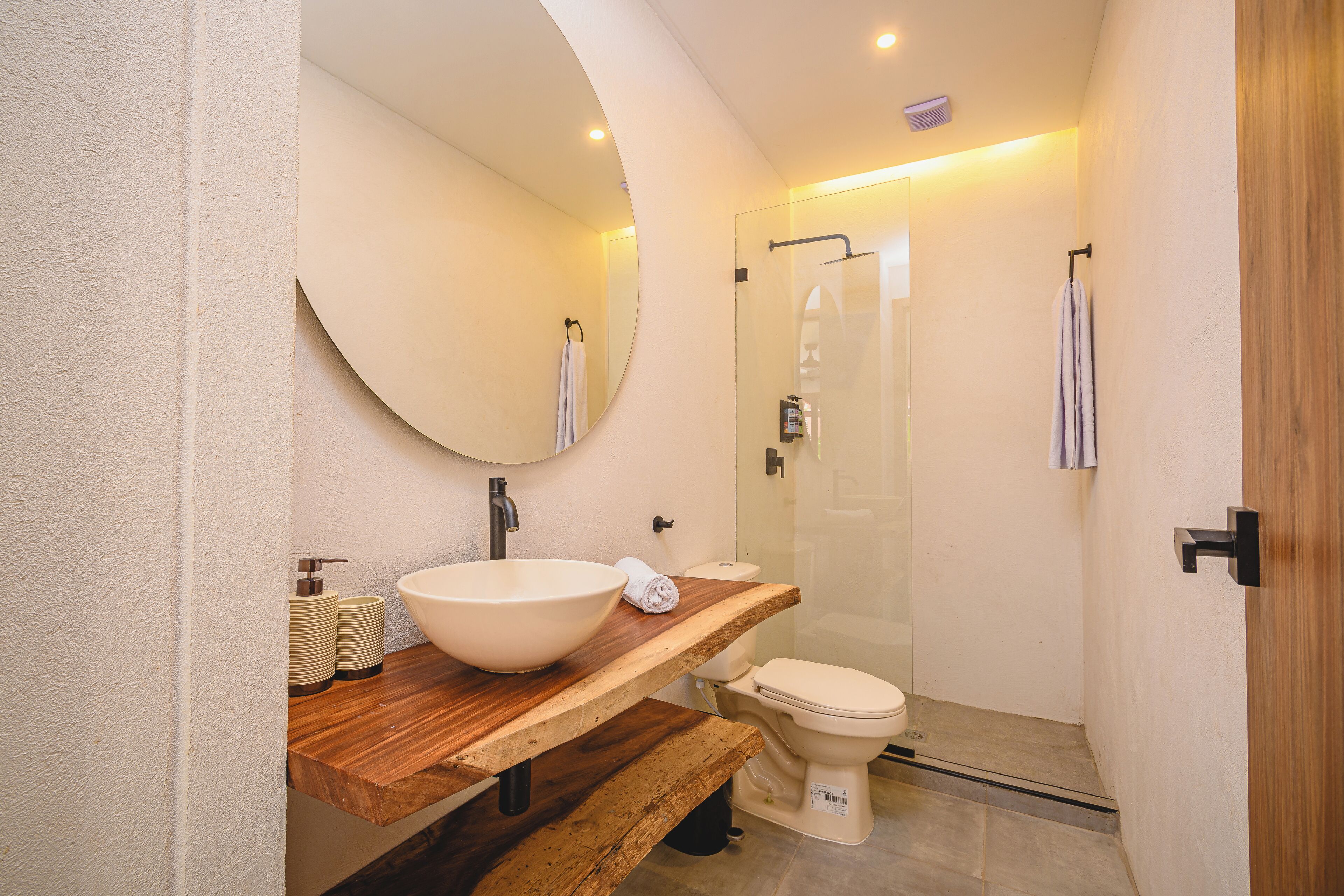 Un baño moderno con un tocador de madera, un espejo redondo grande, un lavamanos blanco y una ducha con cerramiento de vidrio.