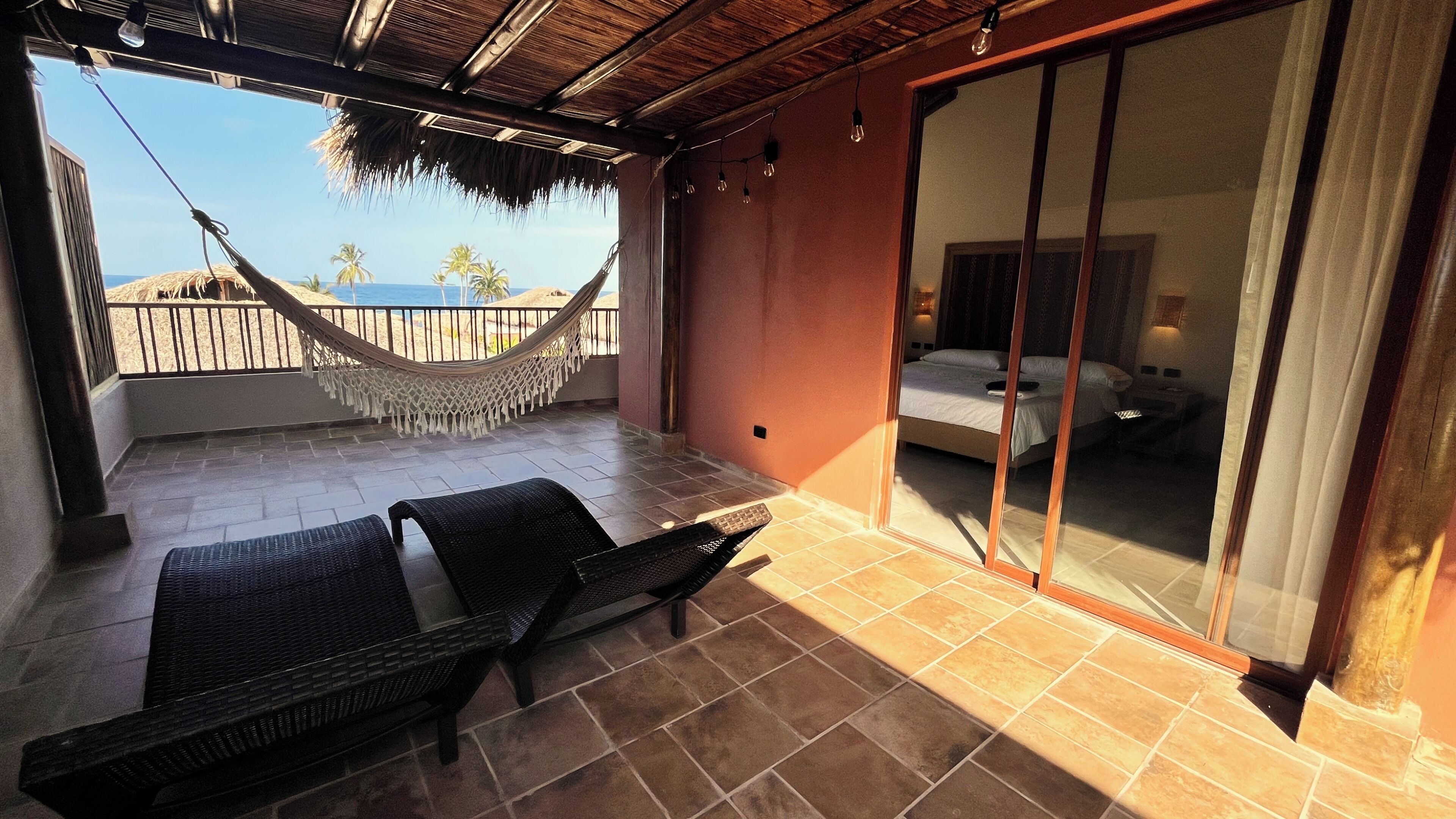 A balcony with a hammock, two wicker chairs, and a bed with a sliding glass door.
