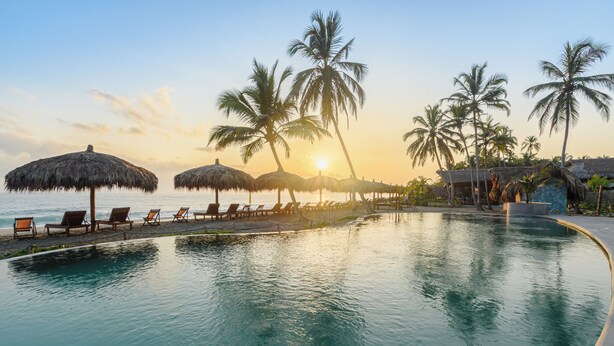 Un espace aménagé au bord de la piscine, avec des chaises longues et des parasols en chaume, entouré de palmiers et offrant une vue imprenable sur le coucher du soleil.
