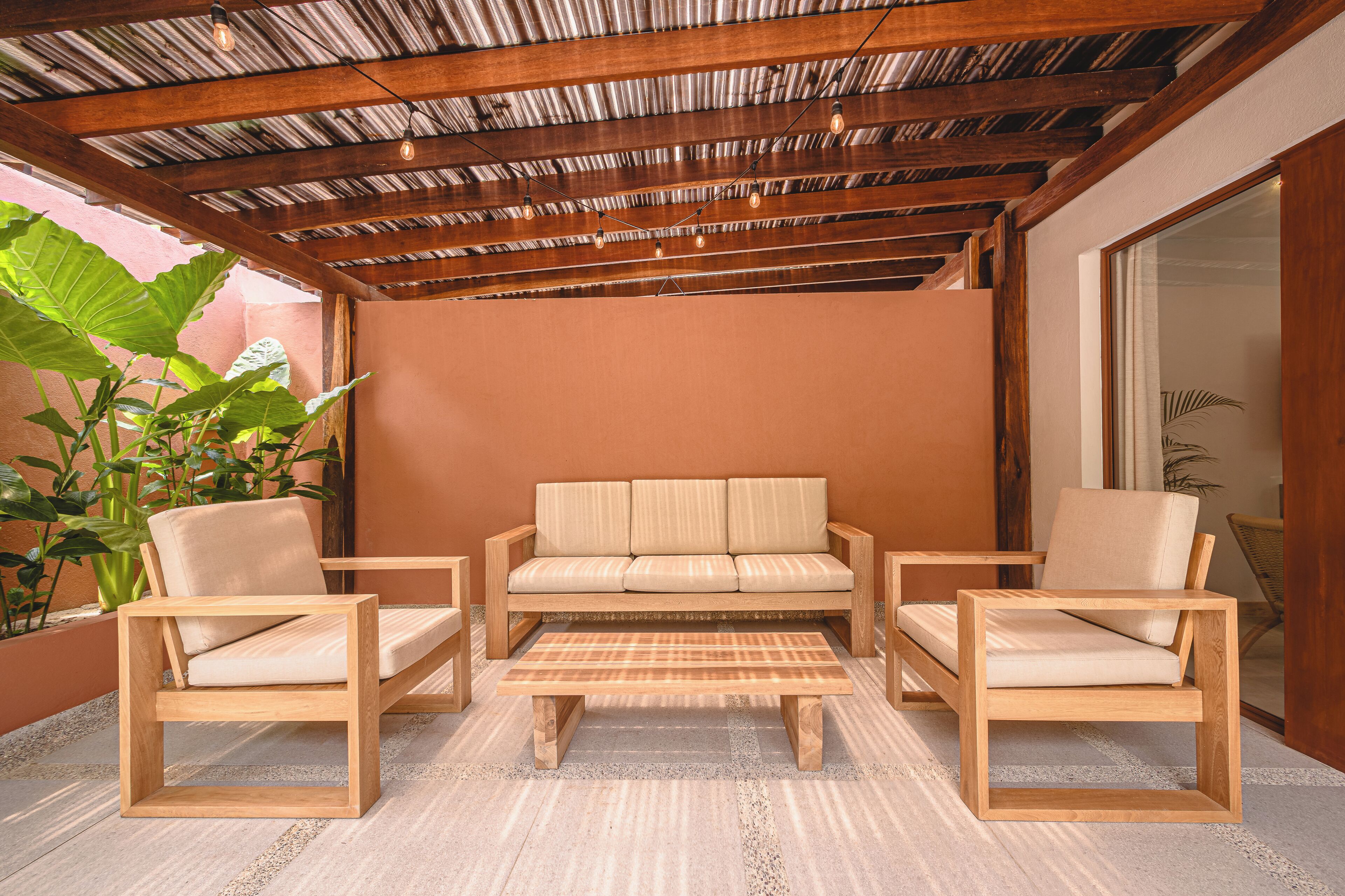 Uma área de estar ao ar livre com bancos de madeira, incluindo um sofá e duas cadeiras, uma mesa de centro, teto com vigas aparentes e luminárias pendentes.