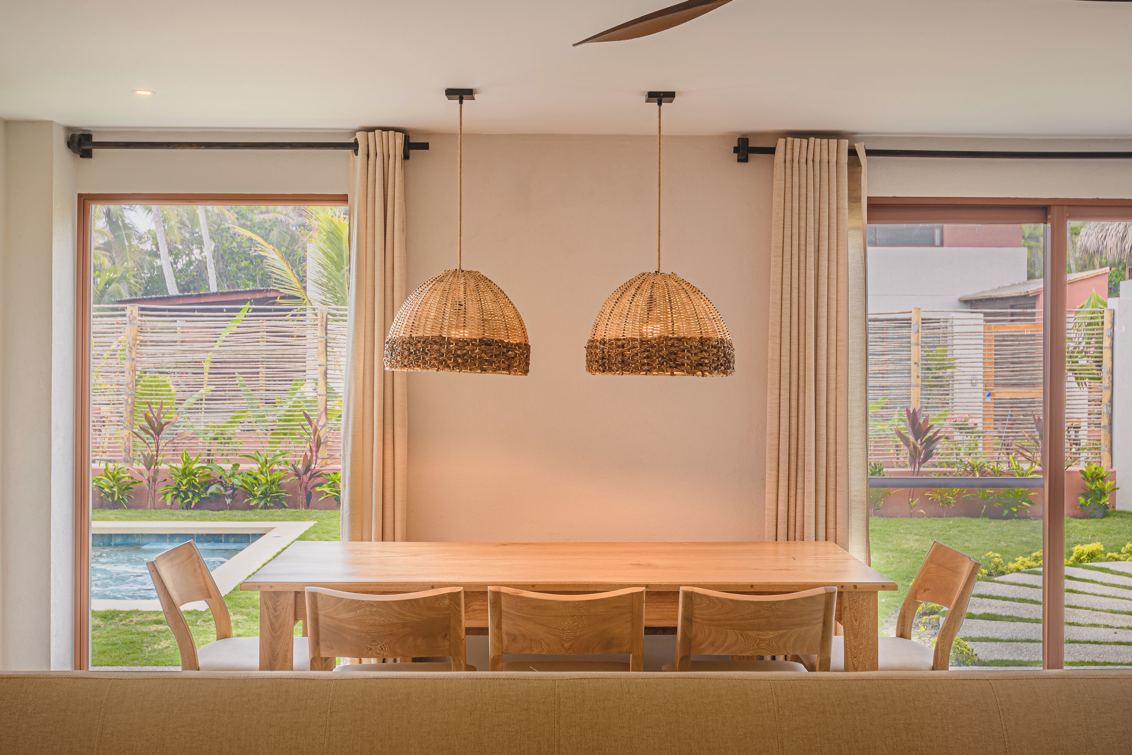 Un coin repas avec une table et des chaises en bois, deux lampes suspendues en osier, et une vue sur une piscine et un jardin, le tout visible à travers de grandes fenêtres.