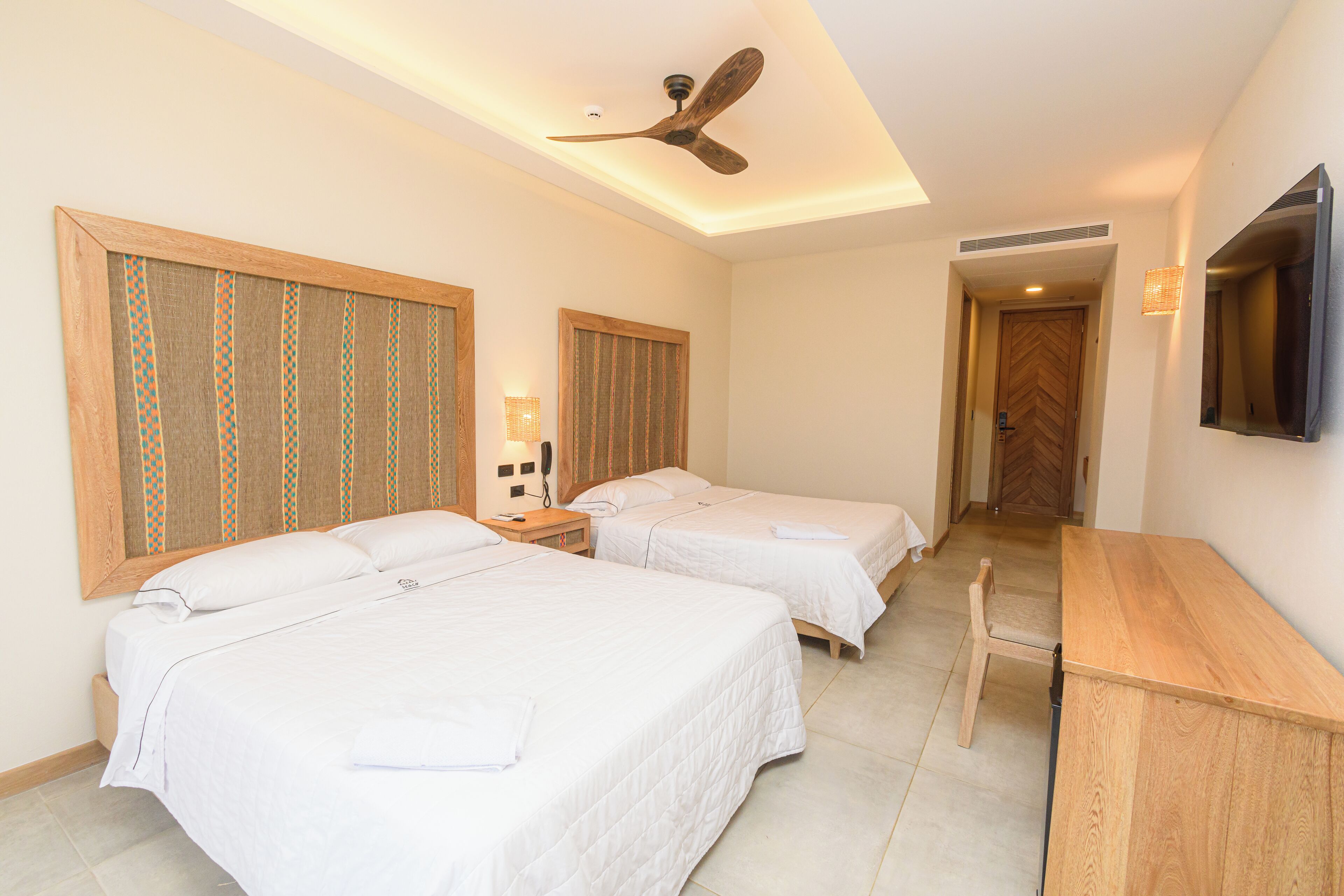 A hotel room with two beds, a desk, a ceiling fan, and a television.