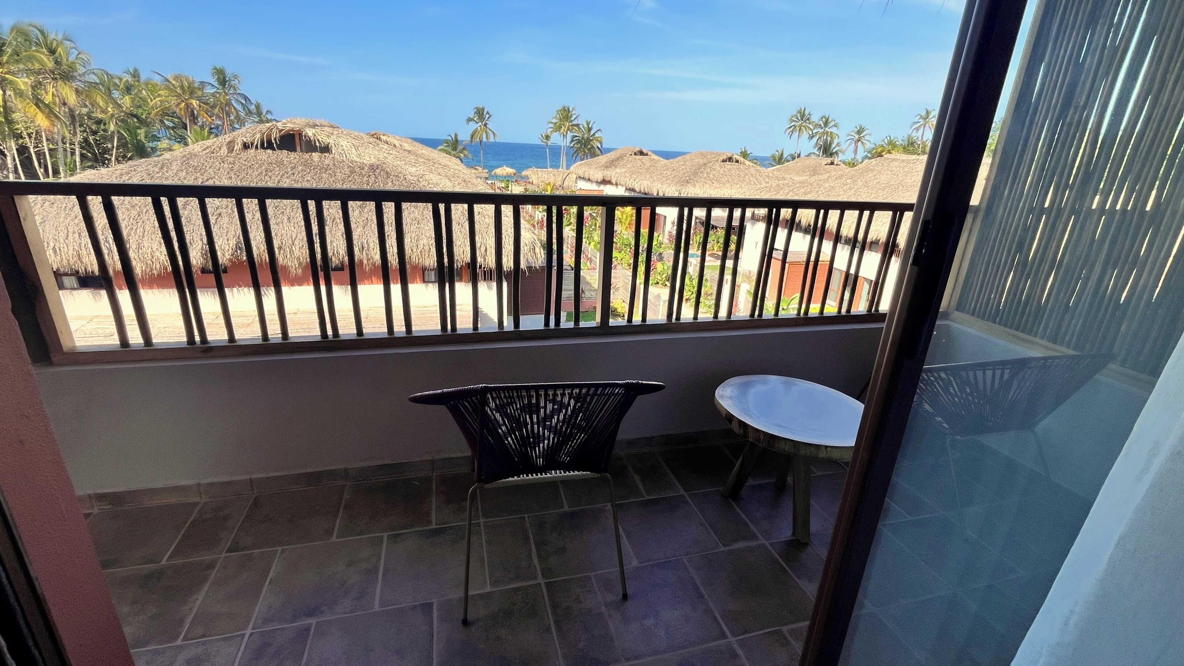 Un balcone con vista sulla spiaggia, una sedia, un tavolino e ombrelloni di paglia.