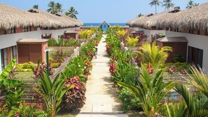 Un sentier bordé de bâtiments aux toits de chaume et d’une végétation tropicale luxuriante, qui mène à une plage.