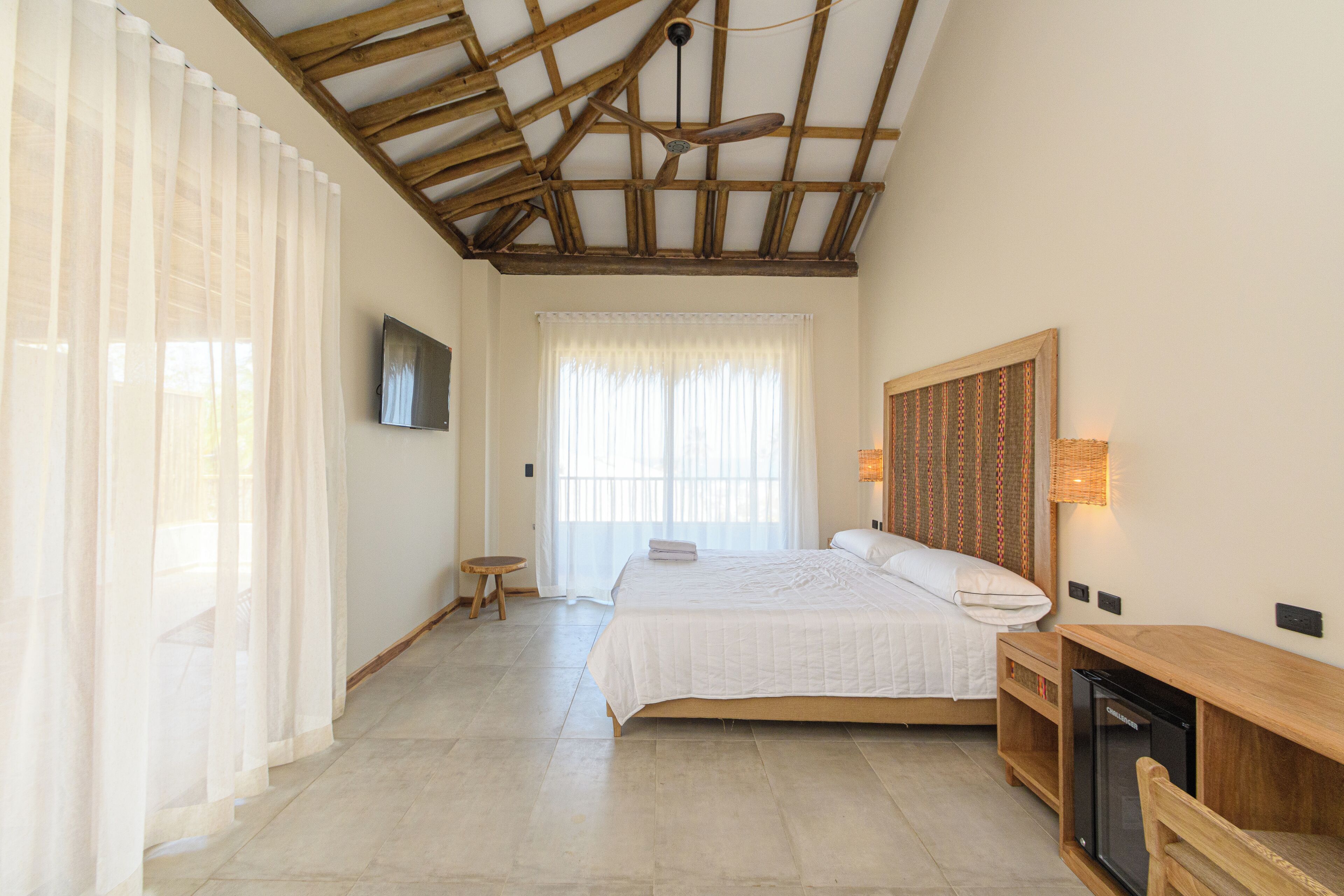 A bedroom with a wooden ceiling, a bed, a TV, a desk, and a window with sheer curtains.