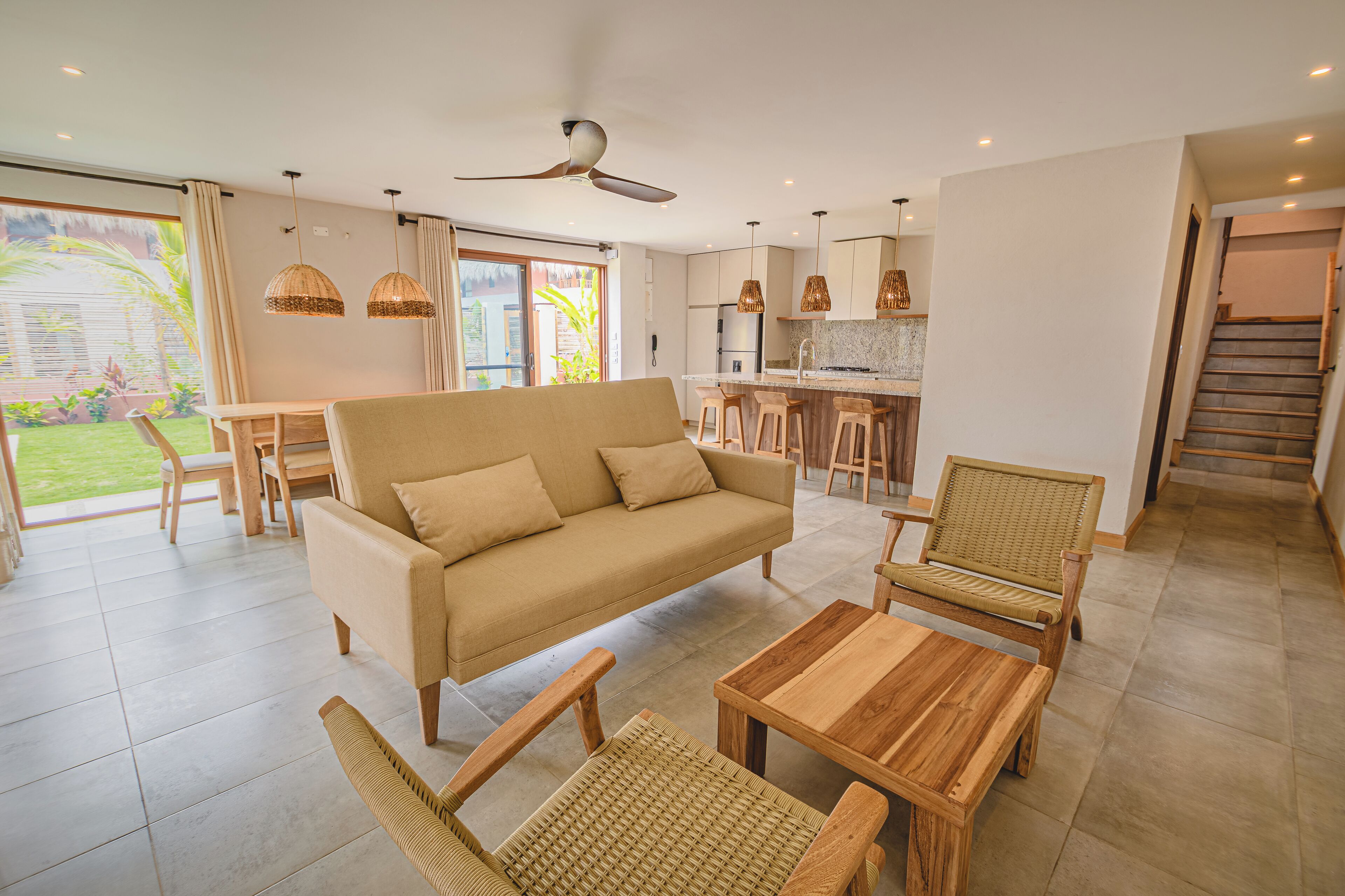 Een moderne woonkamer met een bank, houten stoelen en een salontafel. Er is een keukenhoek met barstoelen en hanglampen.
