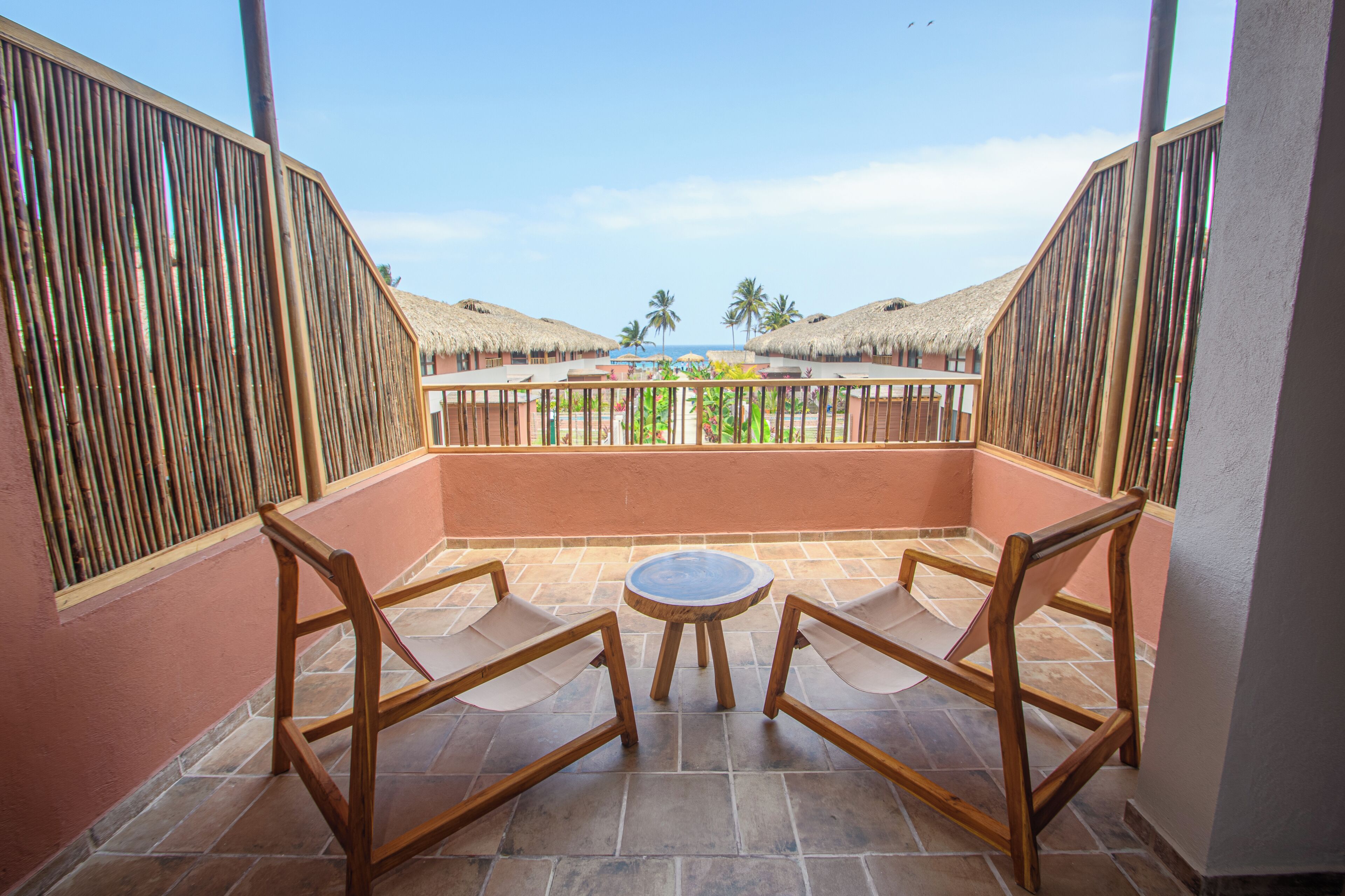 A balcony with two wooden chairs and a small round table, overlooking a beach with thatched-roof huts and palm trees.