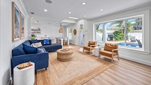 Un salon spacieux avec un canapé bleu, un pouf rond et une table basse en bois. On y trouve également un coin repas avec une table et des chaises, et une vue sur une piscine grâce à de grandes fenêtres.