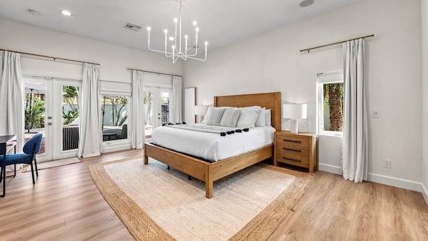 Une chambre spacieuse avec un lit en bois, un lustre, une table de chevet et une vue sur l’extérieur grâce à de grandes fenêtres.