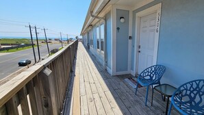 Une terrasse en bois offrant une vue sur la route et l’océan, une porte blanche portant le numéro 5306, et deux chaises bleues.