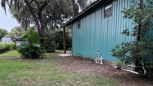 Un bâtiment métallique vert, doté d’une fenêtre et d’une porte, entouré d’une cour verdoyante avec un arbre et un arbuste.