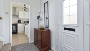 Un couloir mène à une cuisine dotée de meubles blancs, d’un meuble en bois avec un miroir et d’une fenêtre offrant une vue agréable.
