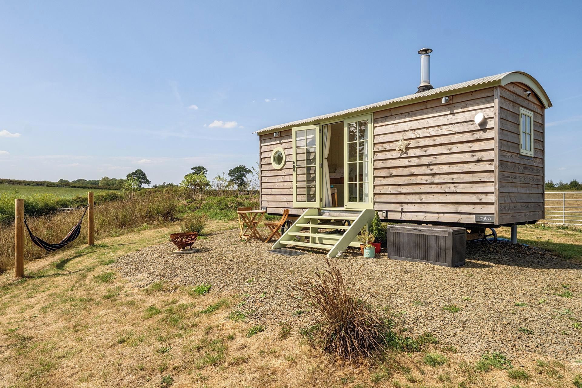 Une cabane en bois avec une véranda, une cheminée et un petit jardin agrémenté d’un hamac et d’un foyer.
