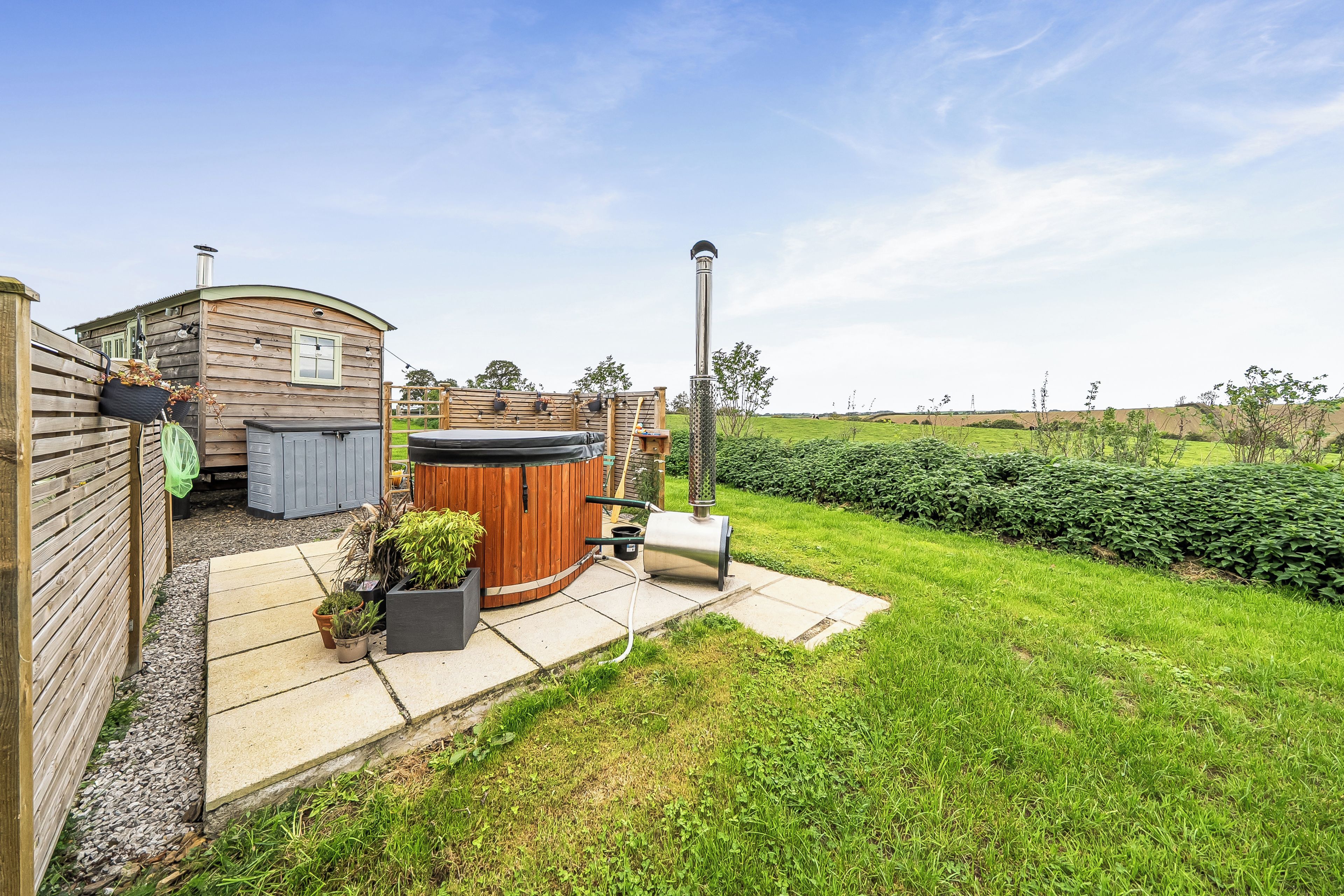 A wooden cabin with a hot tub, a barbecue grill, and a garden area with a lawn and hedges.