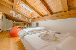 Een slaapkamer met een houten plafond, een bed met witte lakens, een rode poef en een raam.