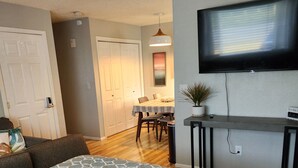 A bedroom with a bed, a TV mounted on the wall, a desk, and a dining area in the background. - Hale Ali'i (Anchorage)