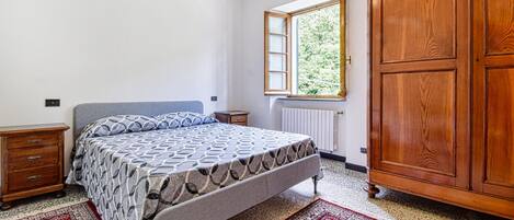 A bedroom with a bed, wooden wardrobe, and a window with a view of greenery.