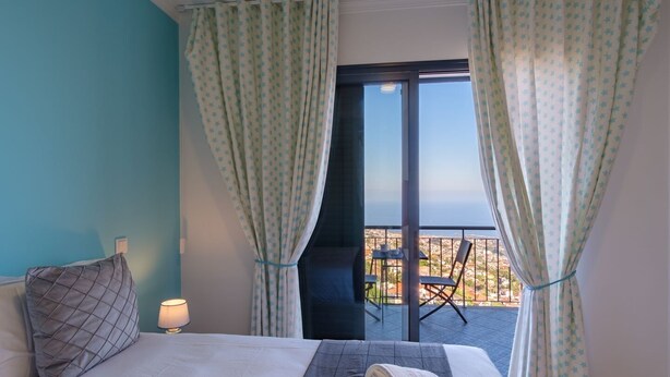 Une chambre d’hôtel avec un lit, une table de chevet, une lampe, une vue sur la ville grâce à une grande fenêtre et un balcon avec une chaise.