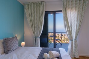 Quarto de hotel com cama, mesa de cabeceira, luminária, vista da cidade por uma janela ampla e varanda com uma cadeira.