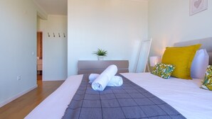Un lit bien fait, avec une couverture grise, une petite plante en pot et un coussin jaune posé sur une tête de lit blanche.