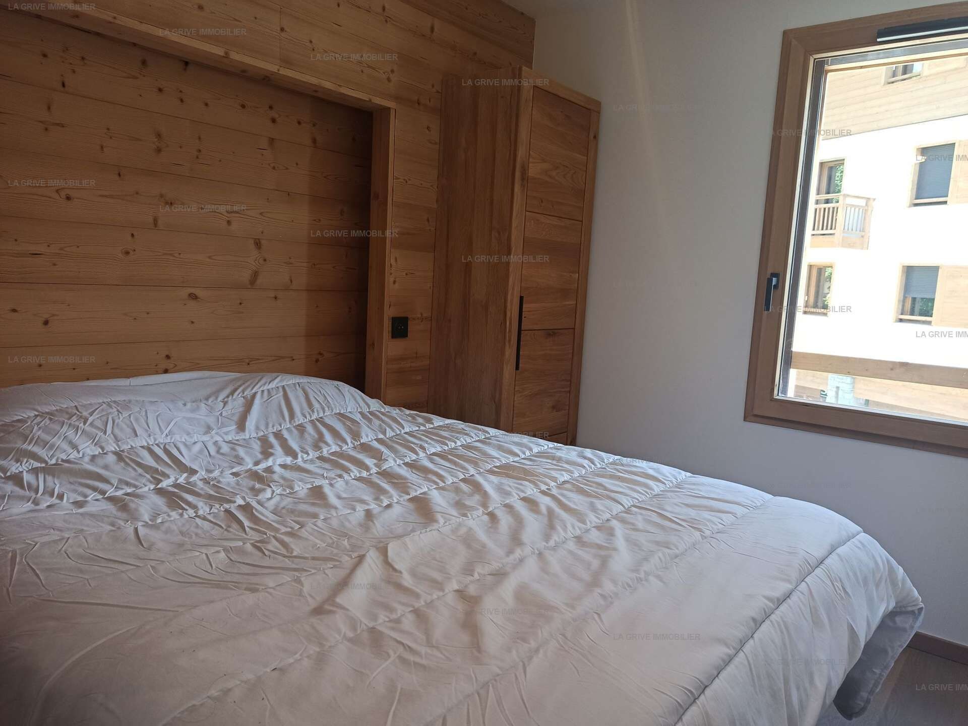 A bedroom with a wooden headboard, a large bed with a white duvet, and a window with a view of buildings.