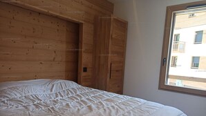 Une chambre à coucher avec une tête de lit en bois, un grand lit recouvert d’une couette blanche et une fenêtre donnant sur des immeubles.
