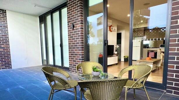 A modern patio with wicker chairs and a glass table, a brick wall, and a sliding glass door leading to a kitchen and living area.