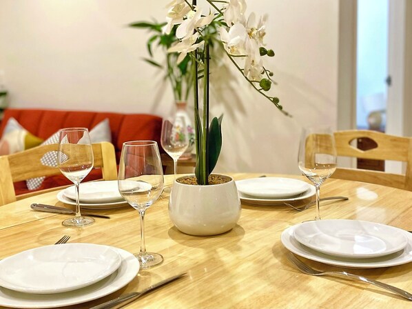 Mesa de comedor dispuesta con platos blancos, copas de vino y cubiertos, con una orquídea en maceta en el centro.