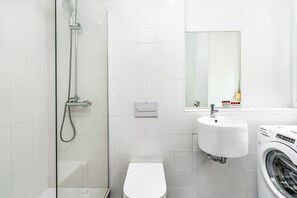Ett modernt badrum med en dusch som är avskärmad med glasväggar, en vit toalett, ett handfat och en tvättmaskin.