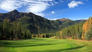 A golf course with a flagstick in the center, surrounded by dense forests and mountains in the background.