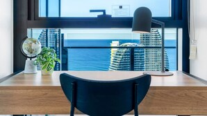 Une chambre moderne avec un bureau en bois, un globe terrestre, une plante en pot et une vue sur l’océan et les immeubles de grande hauteur.