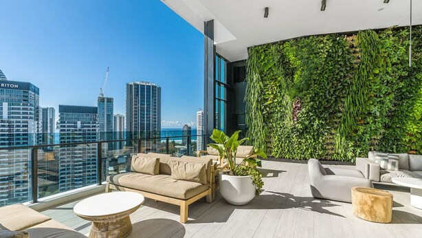 Un salon moderne situé sur le toit, offrant une vue sur la ville, avec un mur végétal, des sièges confortables et une grande plante en pot.