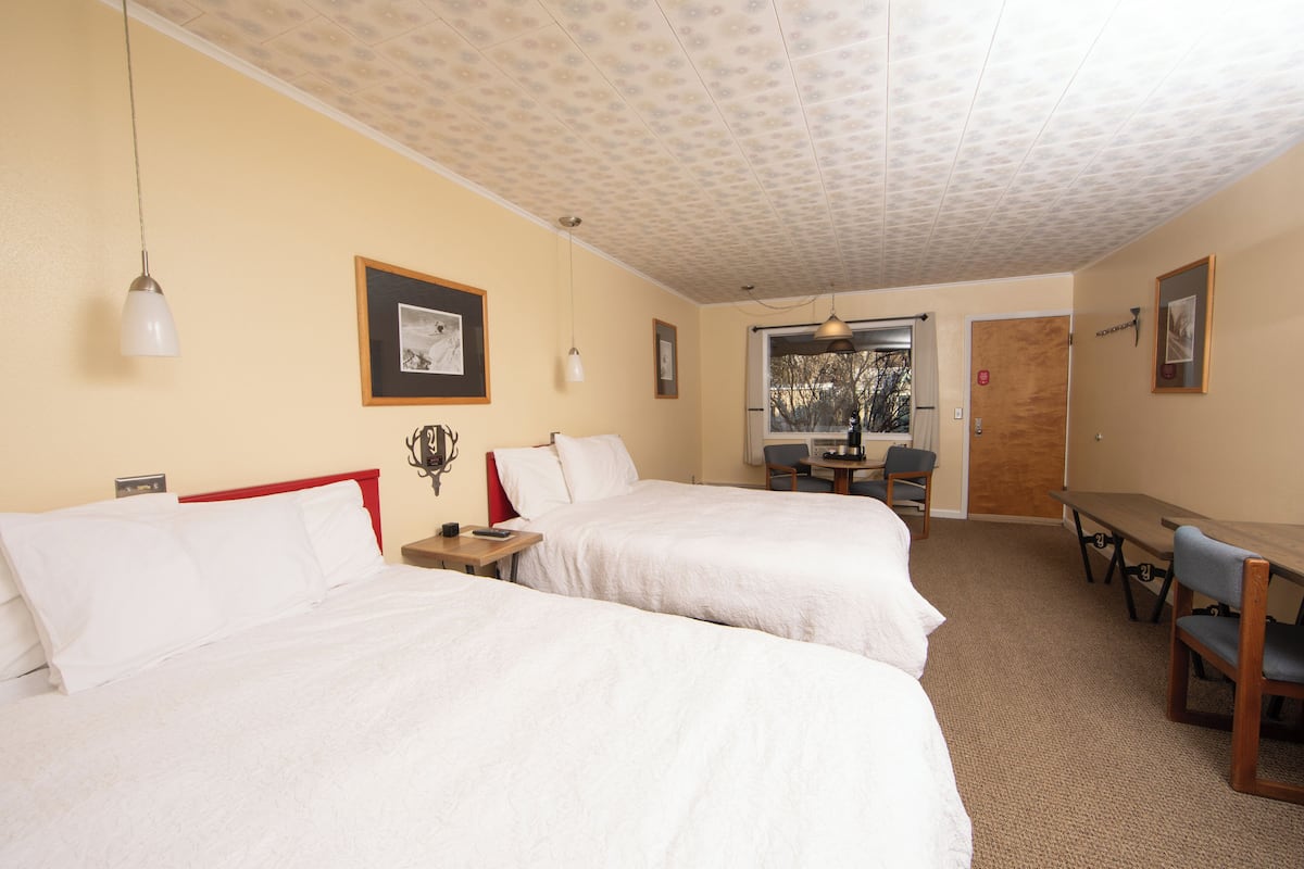 A hotel room with two beds, a wooden table, a chair, and a view of the outdoors.