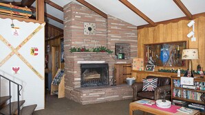 A cozy living room with a brick fireplace, wooden beams, a bookshelf, and a leather armchair. - Yodeler Motel (Red Lodge)