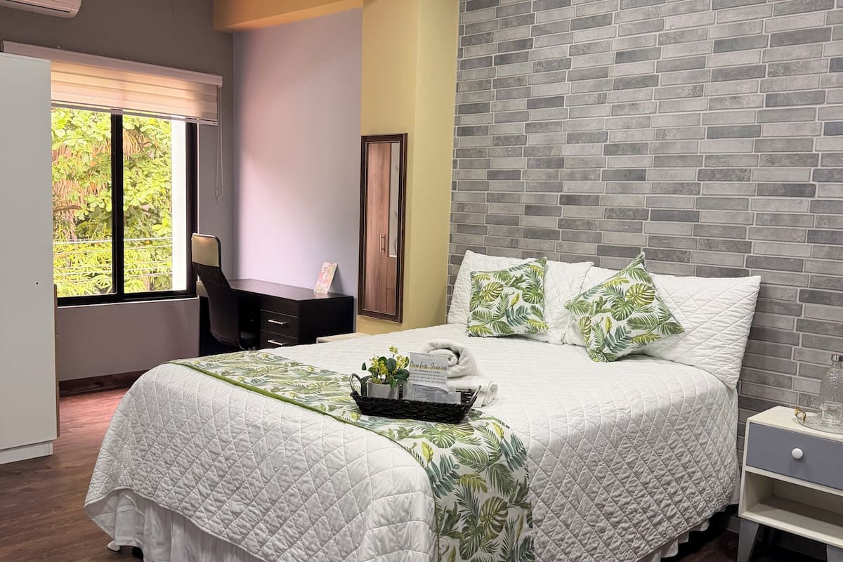 A bedroom with a bed, a nightstand, a desk, and a window with a view of greenery.