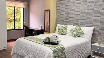 A bedroom with a bed, a nightstand, a desk, and a window with a view of greenery.