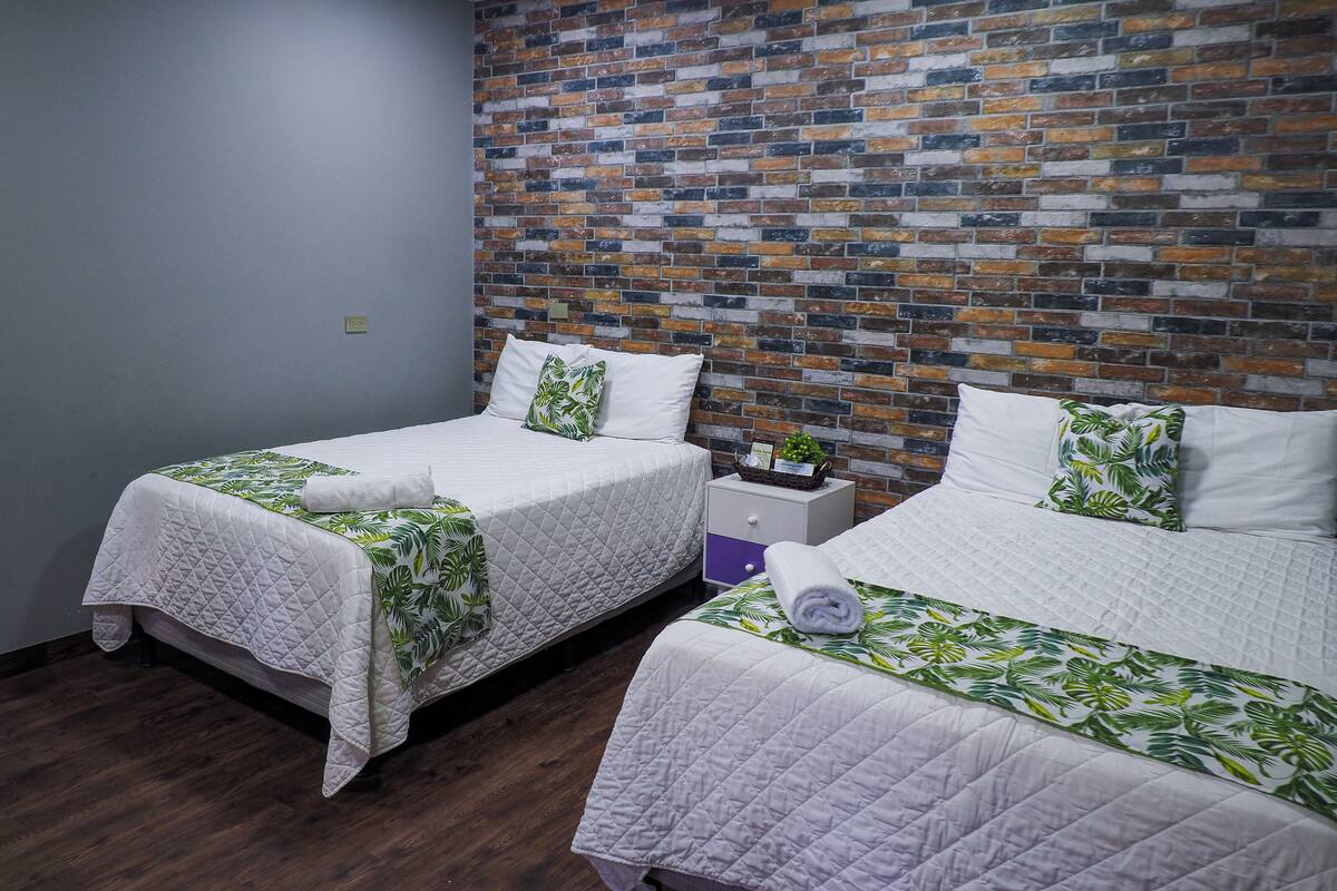A hotel room with two single beds, each with a white quilted bedspread and green leaf-patterned pillows. The room features a brick-patterned wall and wooden flooring.