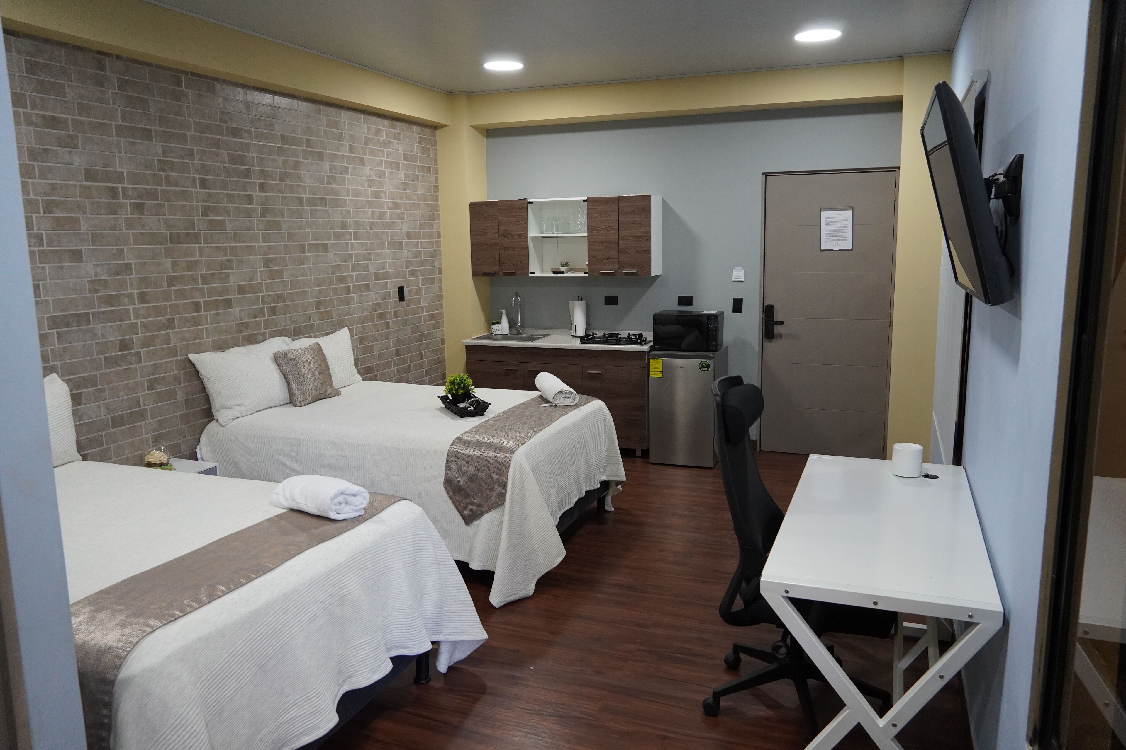 A hotel room with two beds, a small kitchenette, a desk, and a TV.