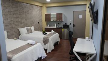 A hotel room with two beds, a small kitchenette, a desk, and a TV.