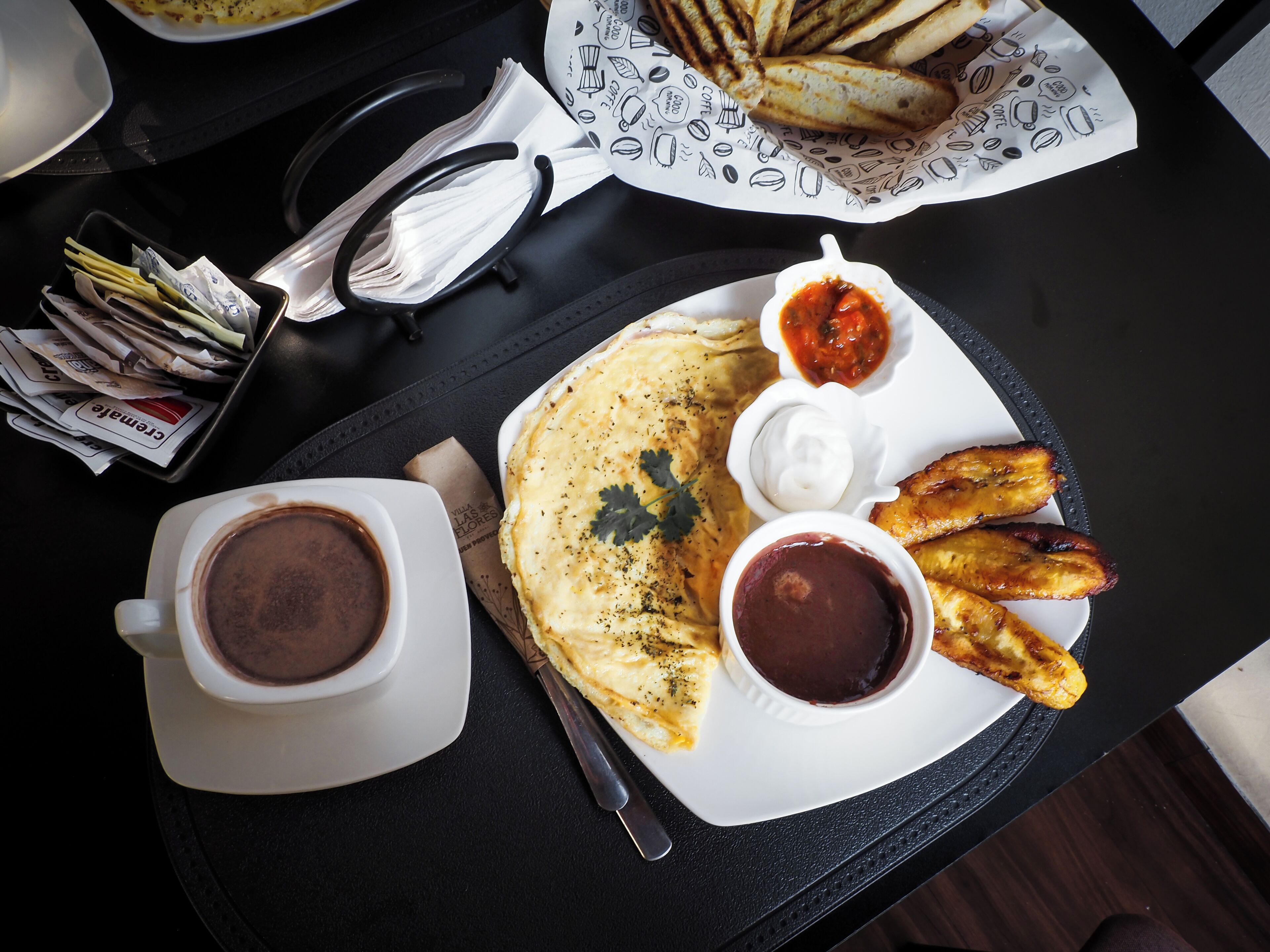 Un plato de desayuno con tortitas, plátanos fritos y dos salsas, acompañado de una taza de café.