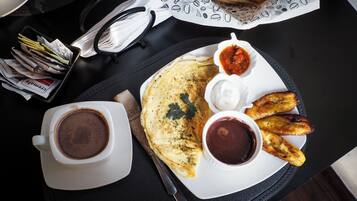 Un plato de desayuno con tortitas, plátanos fritos y dos salsas, acompañado de una taza de café.