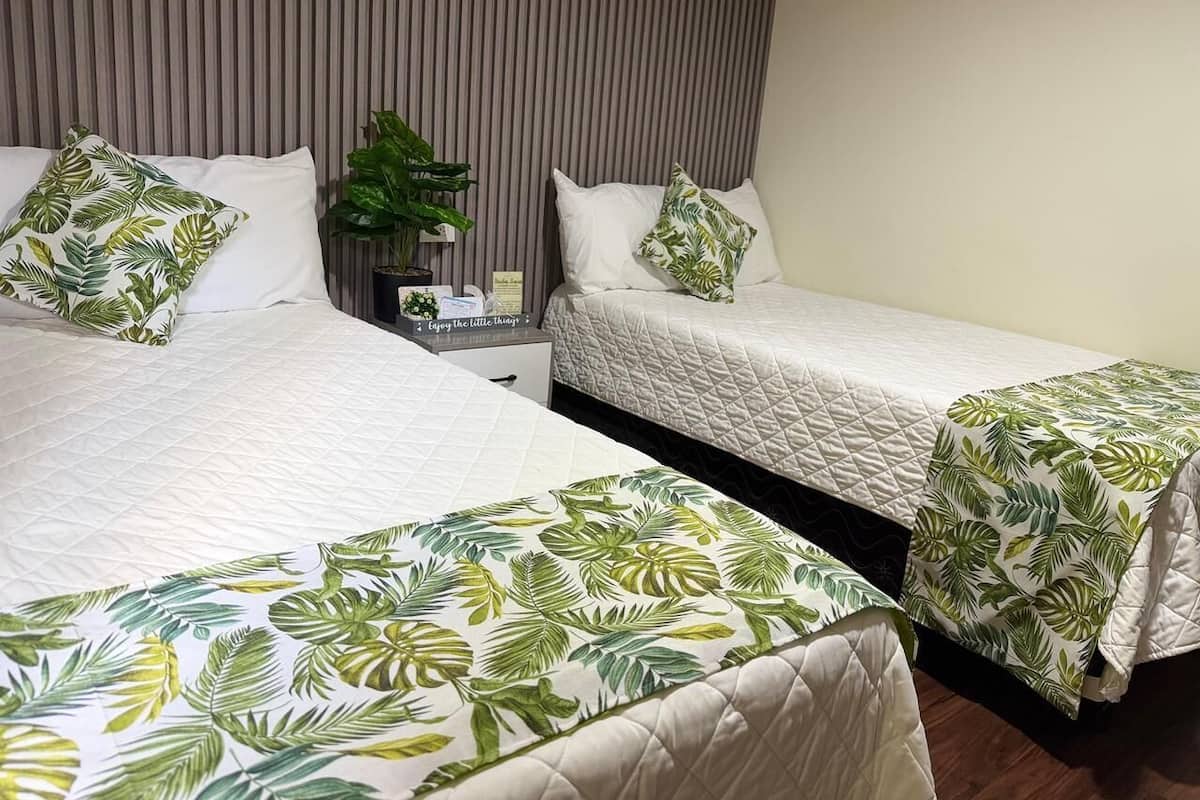 A hotel room with two beds, each with a white quilted bedspread and a tropical leaf-patterned bed skirt. There is a nightstand with a small potted plant and a sign that reads "Enjoy the little things."