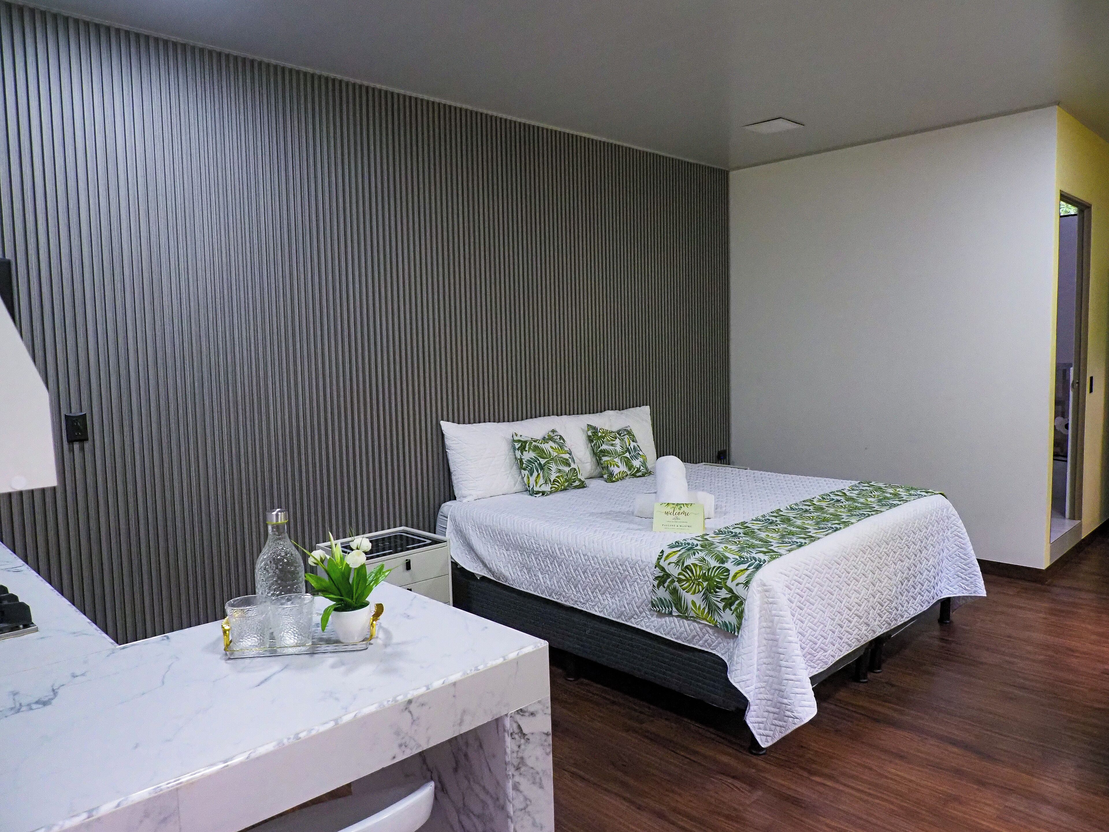 Ein modernes Hotelzimmer mit einem Bett, einem Nachttisch mit einem Glas, einer Pflanze und einem Willkommensschild.