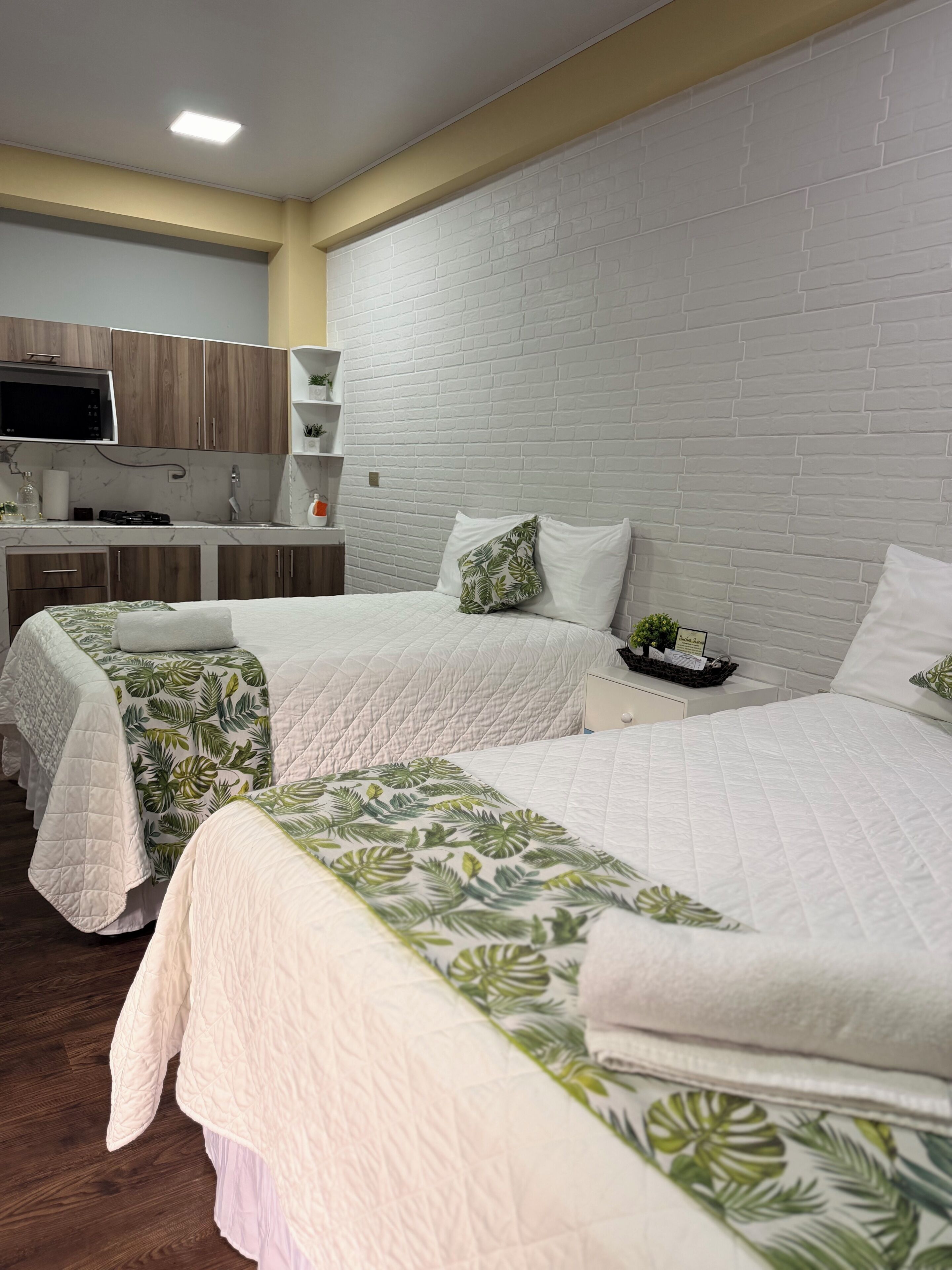 A hotel room with two beds, a kitchenette, a white brick wall, and a television.