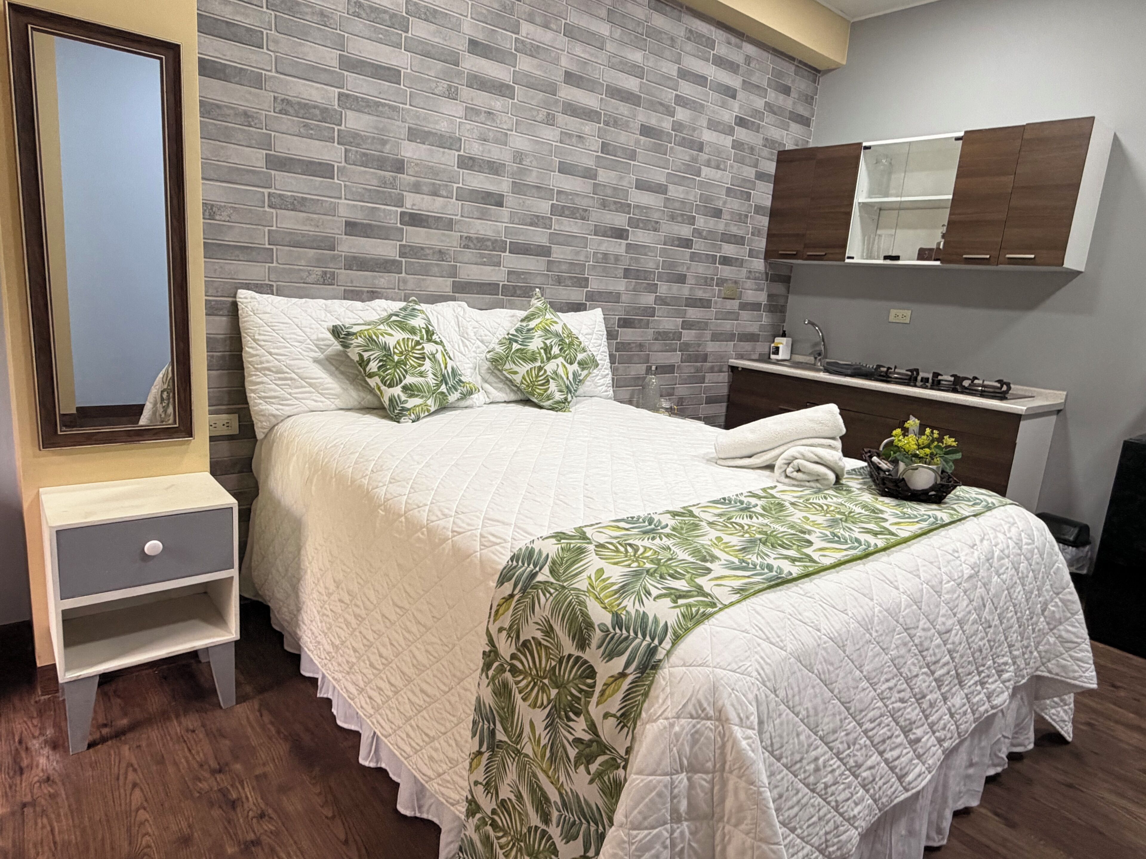 Une chambre avec un mur décoré d’un motif en briques, un lit recouvert d’une courtepointe blanche, deux oreillers, une table de chevet avec un tiroir, une petite table d’appoint, un miroir, une petite plante et un meuble fixé au mur.