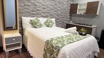 Une chambre avec un mur décoré d’un motif en briques, un lit recouvert d’une courtepointe blanche, deux oreillers, une table de chevet avec un tiroir, une petite table d’appoint, un miroir, une petite plante et un meuble fixé au mur.