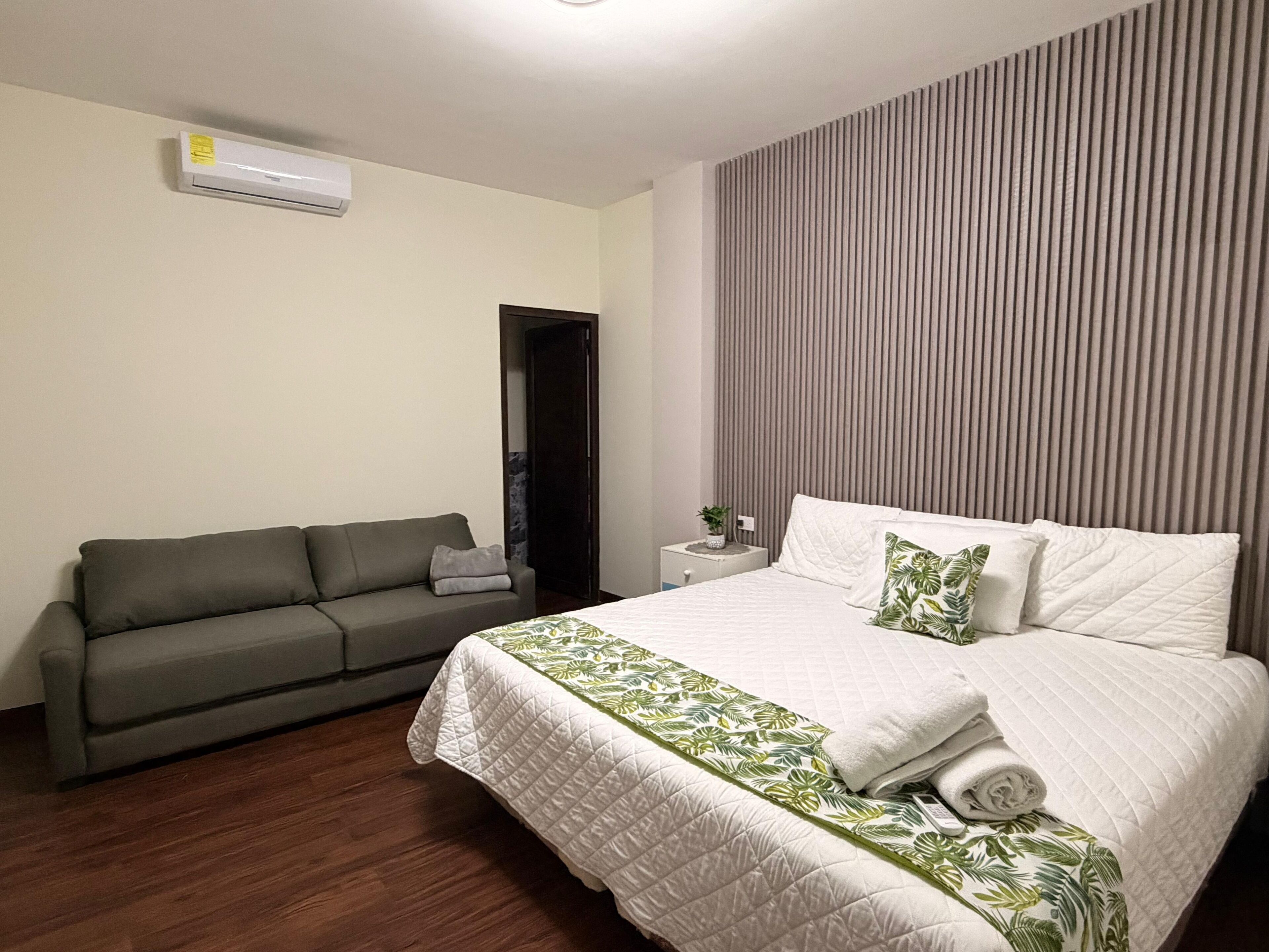 A hotel room with a bed, a grey sofa, a small bedside table with a plant, and a wall-mounted air conditioner.